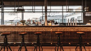A bar area with stools, a bar counter, and a view of a Ferris wheel through large windows. - Superbude Wien Prater (Vienna)