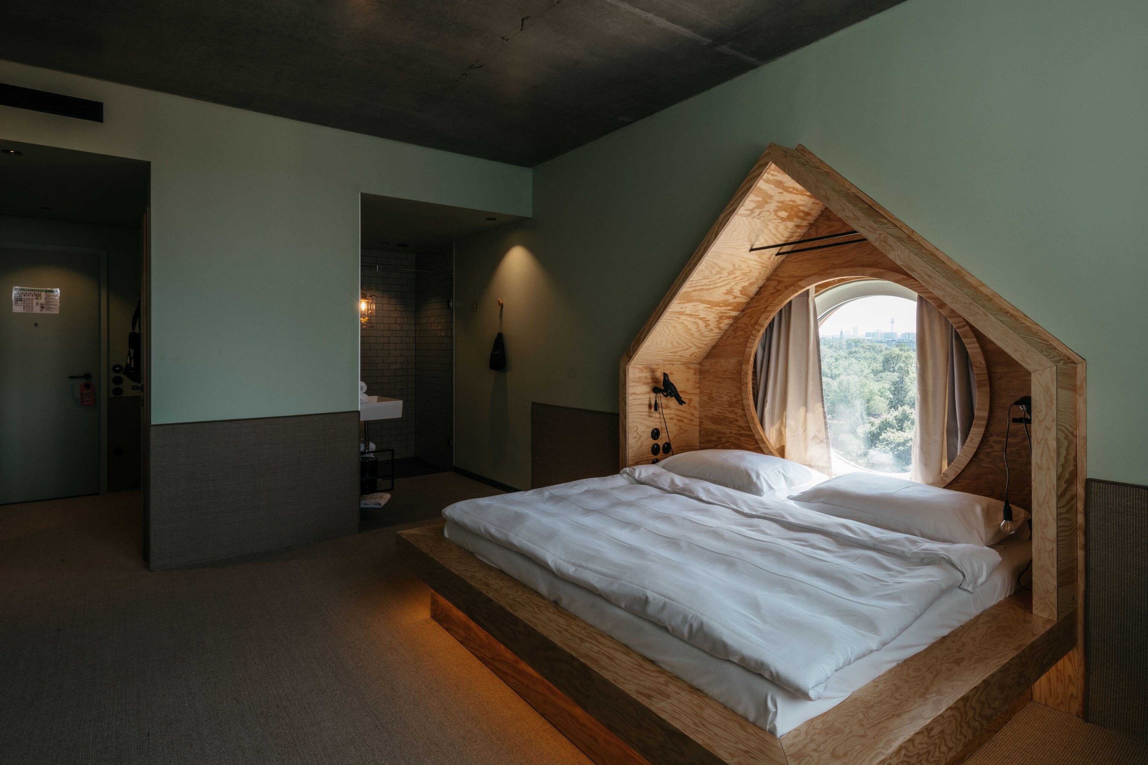 Ein Schlafzimmer mit schrägen Wänden, einem eingebauten Bett, einem kleinen Badezimmer und Blick auf die Stadt durch ein Fenster.