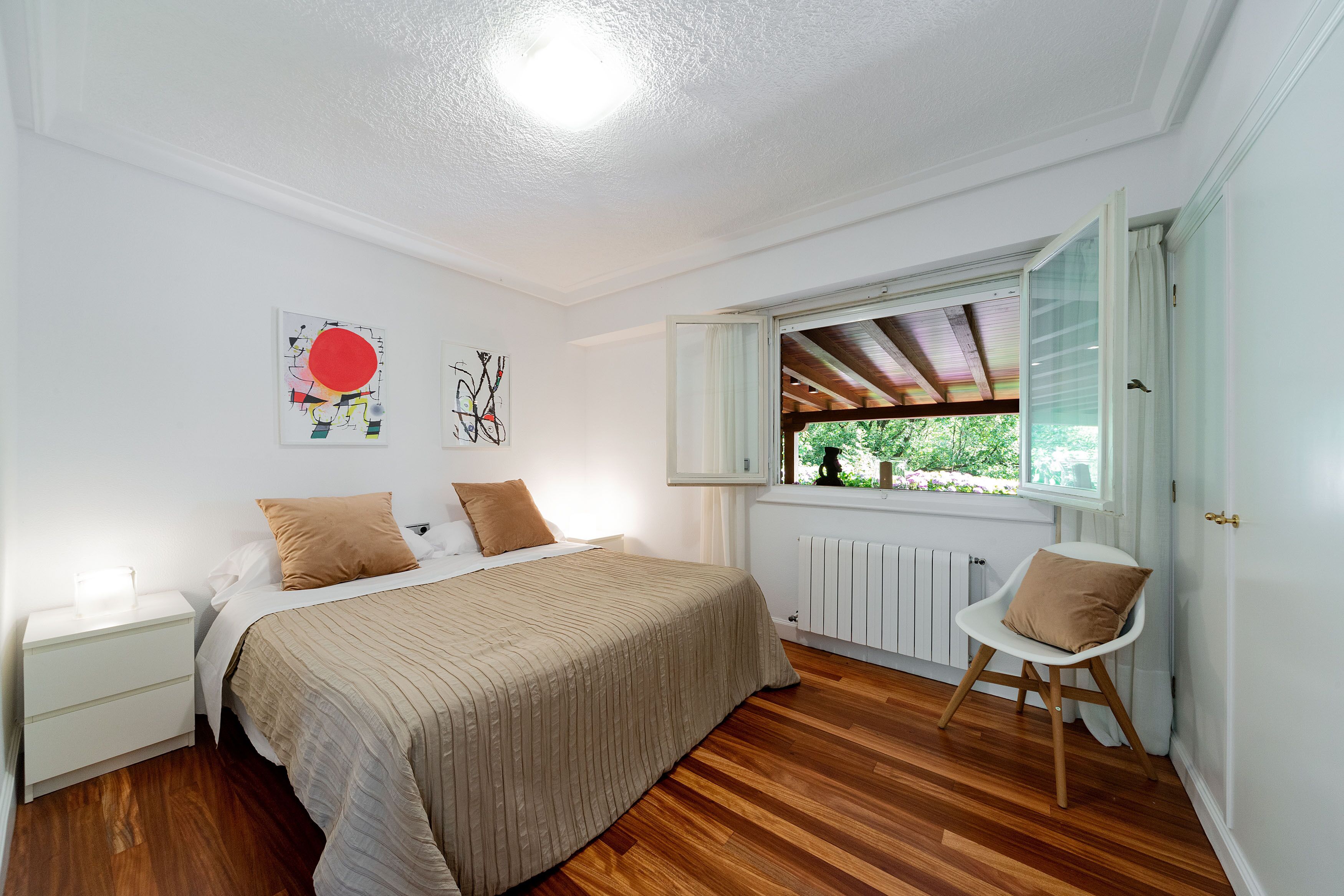 A bedroom with a bed, a nightstand, a chair, a window with a view, and artwork on the wall.