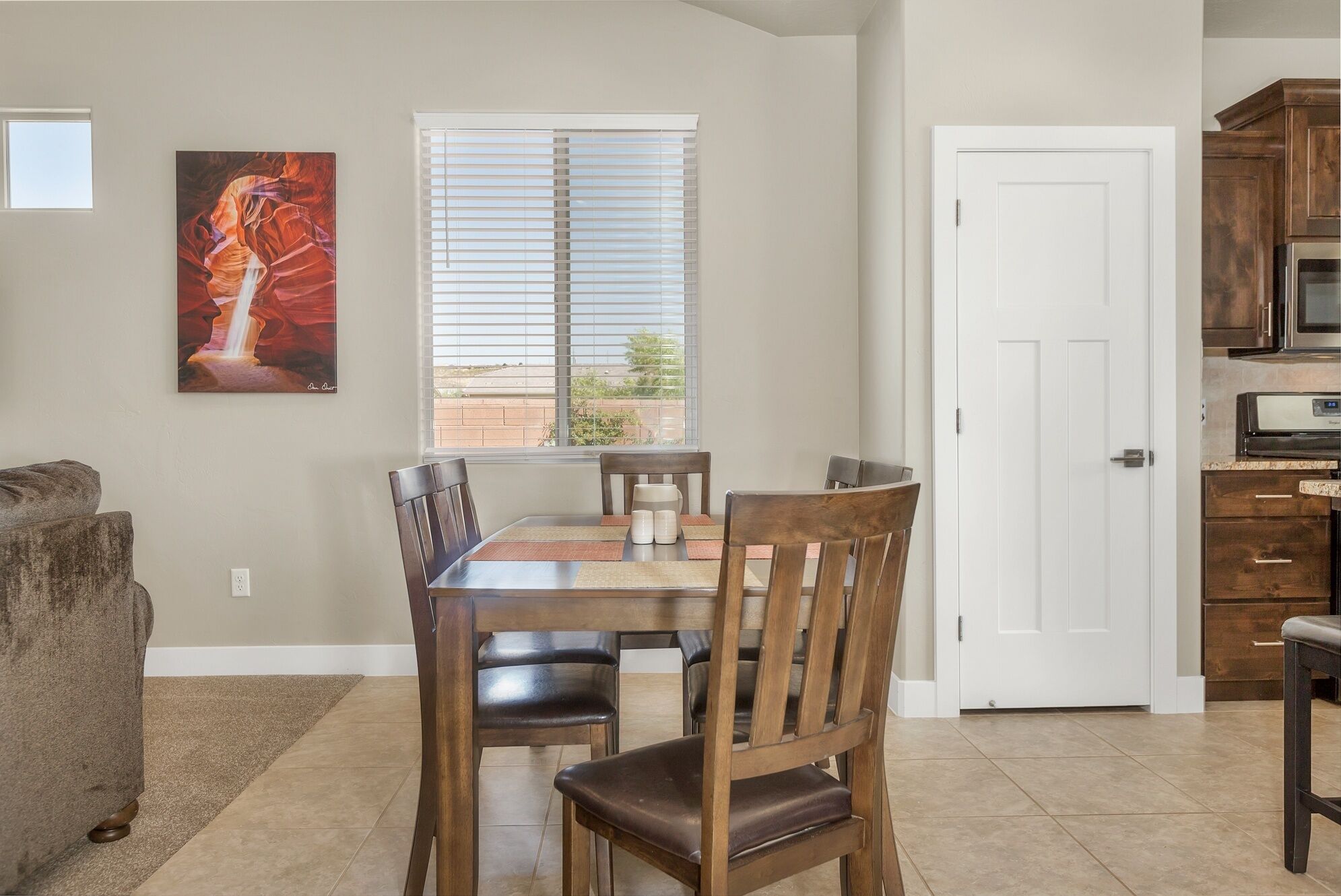 A dining area with a table and chairs, a painting on the wall, a window with blinds, and a kitchen with cabinets and appliances.