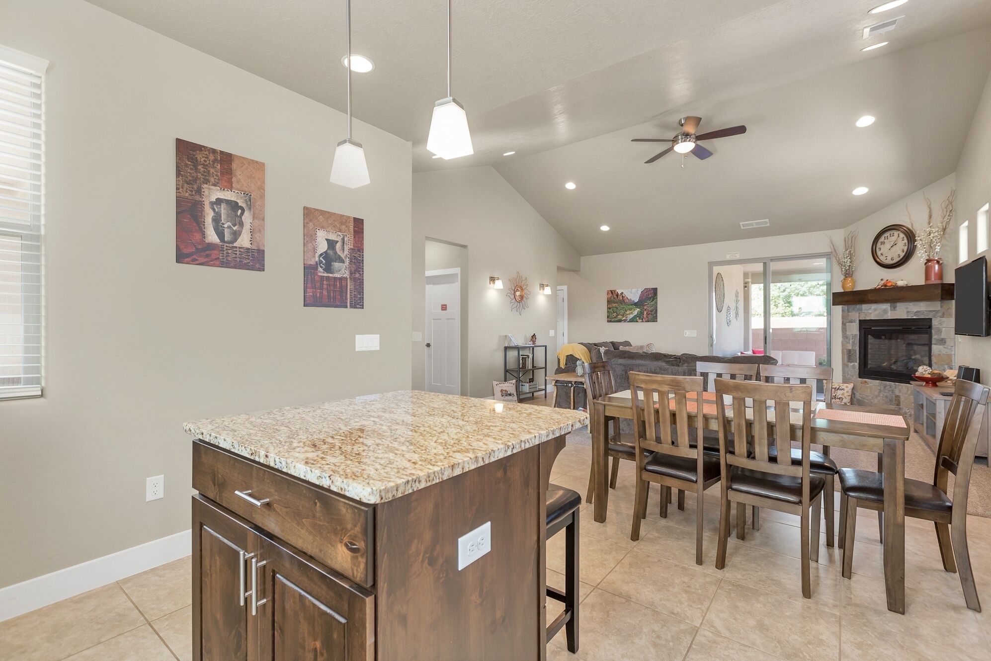 A spacious kitchen with a granite island, wooden dining table, and chairs. There is a ceiling fan, recessed lighting, and artwork on the walls.