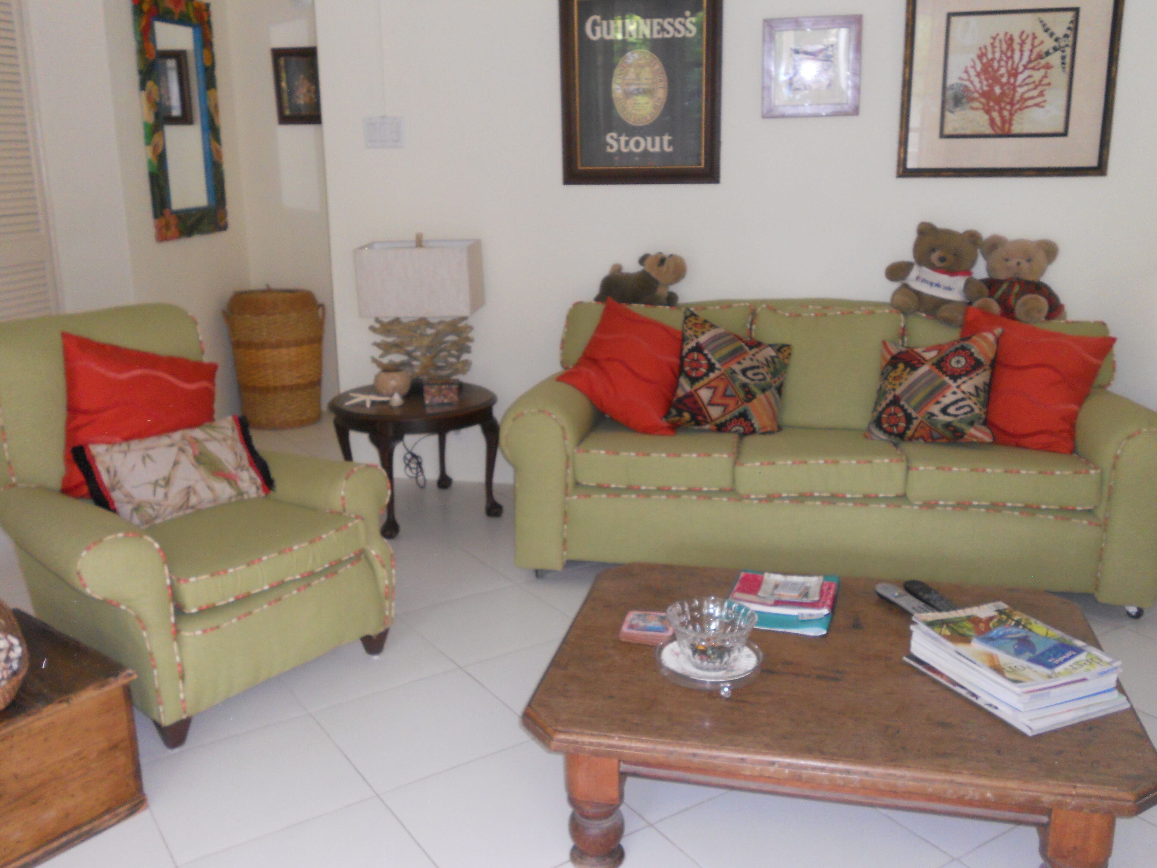 Un salon avec un canapé vert, une table basse en bois et un fauteuil vert. On peut voir deux ours en peluche sur le canapé, un tableau encadré au mur et une lampe sur une table d’appoint.