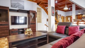 Een moderne woonkamer met een flatscreen televisie, houten meubels, een bank met rode kussens en een trap naar een hoger gelegen verdieping.