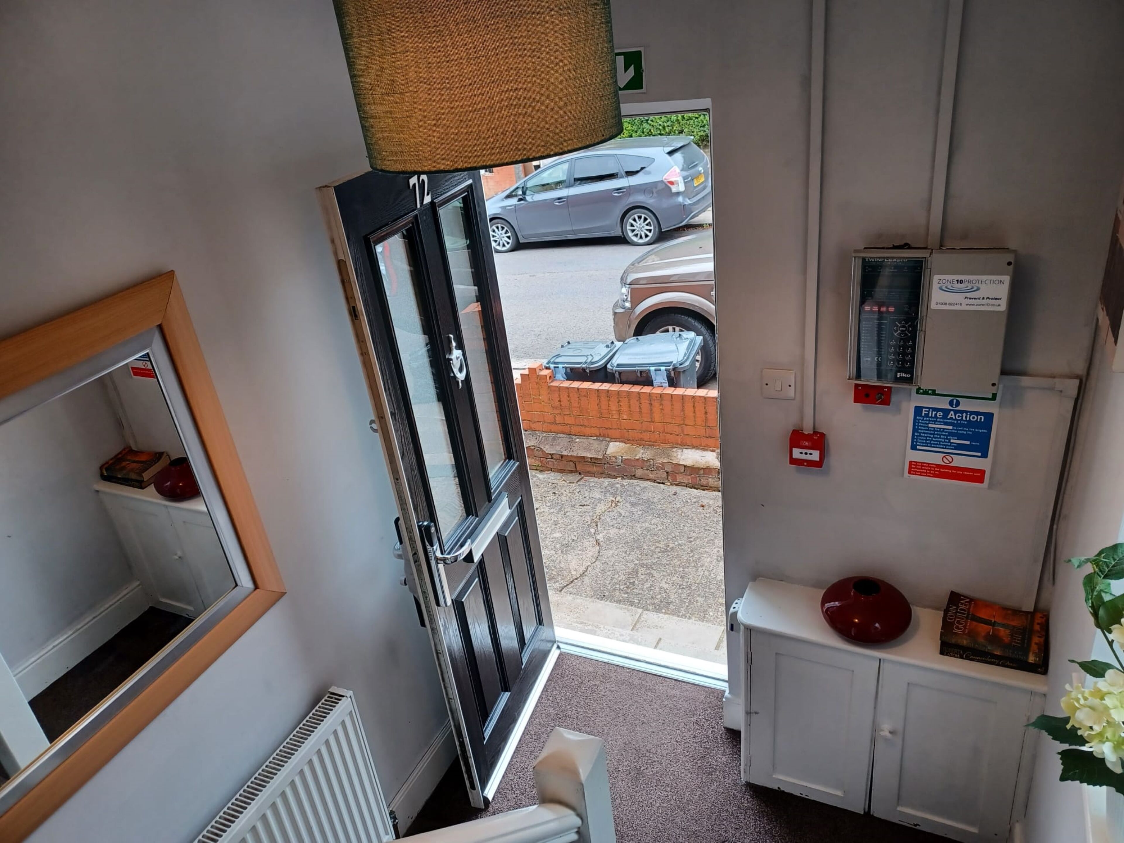 A hotel room with a large window, a radiator, a cabinet with a red vase, and a fire action sign.