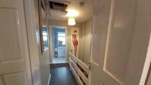 Un pasillo estrecho con una puerta blanca a la derecha, una lámpara arriba y una vista a una cocina con un lavamanos y alacenas.