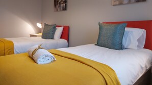 Habitación de hotel con dos camas, cada una con cabecero rojo, ropa de cama blanca y una manta amarilla. Hay dos cuadros colgados en la pared, una lámpara de noche y una mesita con una lámpara.