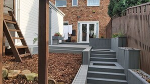 Patio trasero residencial con una escalera de madera, una casa de ladrillo, una valla de madera y un conjunto de escalones grises que conducen a una puerta.