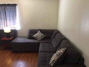 Ein Wohnzimmer mit einem dunkelgrauen Ecksofa, zwei gemusterten Zierkissen, einem kleinen Beistelltisch mit Lampe und einem Fenster mit Vorhängen.