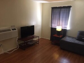 Ein Wohnzimmer mit Flachbildfernseher auf einem Schrank, einer Couch, einem kleinen Tisch mit einer Lampe und einem Fenster mit Vorhängen.