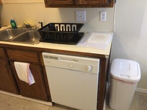 Een keuken met een vaatwasser, een prullenbak en een zwart afdruiprek op het aanrecht.