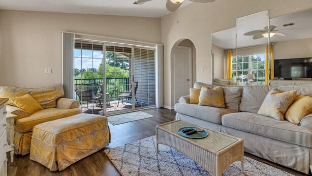 A spacious living room with a sofa, ottoman, and a coffee table. There is a sliding glass door leading to an outdoor patio area.