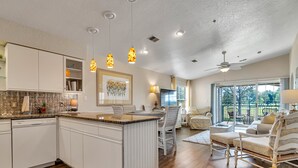 A spacious kitchen with a central island, white cabinetry, and pendant lighting. There is a dining area with a table and chairs, and a living room with a sofa and a view of the outdoors.