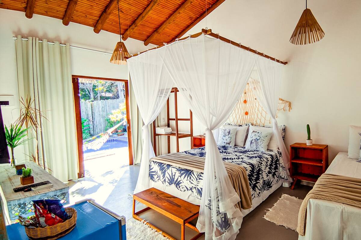 A bedroom with a canopy bed, a bedside table, a nightstand, a small table with a basket, and a view of the outside.