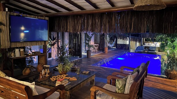 Un área de piscina con mobiliario de madera, un televisor y plantas tropicales.