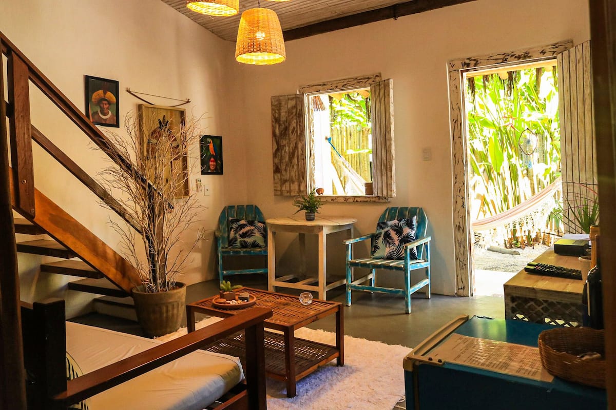 A cozy room with a wooden staircase, a dining table, two blue chairs, a small round table, and a view of a bamboo garden through a window.