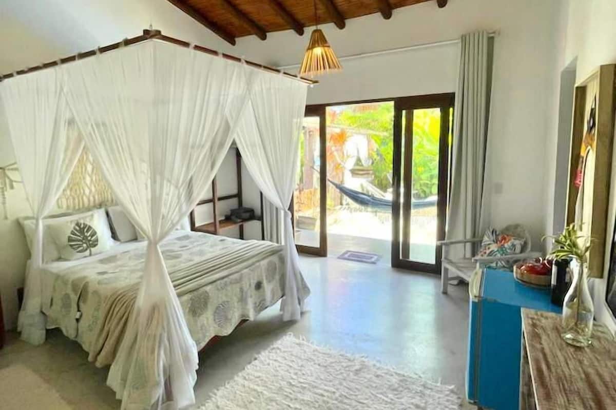A bedroom with a four-poster bed, a wooden ceiling, a large window, and a view of greenery outside.