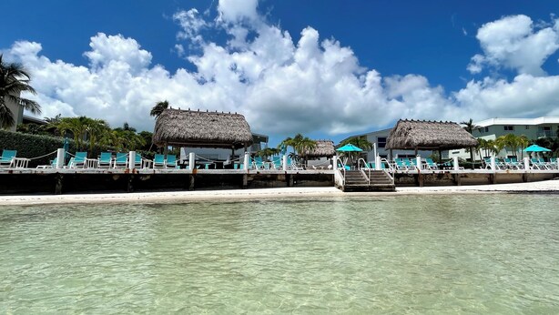 A beachfront resort with thatched-roof structures, clear turquoise water, and a sandy beach. - Beachside Unit 36 - Key Colony (Key Colony Beach)