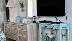 A living room with a large flat-screen TV, a light blue console table, a wicker cabinet, a lamp, and a framed picture of a sea turtle. - Beachside Unit 14 - Key Colony (Key Colony Beach)