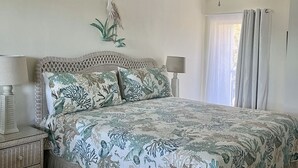 A bedroom with a bed, nightstands, a wall-mounted lamp, and a window with curtains. - Beachside Unit 14 - Key Colony (Key Colony Beach)