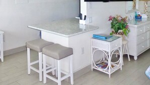 A small, modern kitchen with a flat-screen TV, white bar stools, and a wicker side table with a book and a plant. - Beachside Unit 34 - Key Colony (Key Colony Beach)