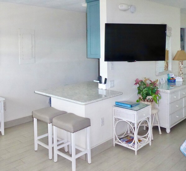 A small, modern kitchen with a flat-screen TV, white bar stools, and a wicker side table with a book and a plant. - Beachside Unit 34 - Key Colony (Key Colony Beach)