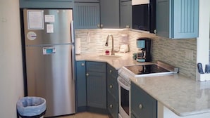 A kitchen with teal cabinets, a stainless steel refrigerator, a microwave, a stove, and a sink. There are two trash bins and a countertop with a coffee maker. - Beachside Unit 34 - Key Colony (Key Colony Beach)