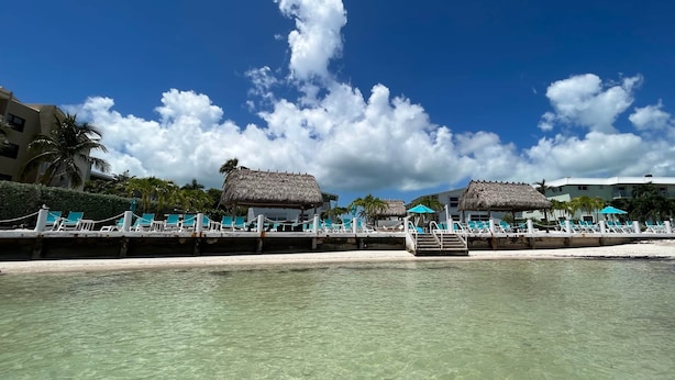Un resort frente al mar con estructuras de techo de paja, agua turquesa cristalina y una playa de arena blanca.