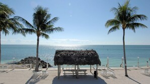 A beach with a thatched hut, palm trees, and lounge chairs. - Beachside Unit 29 - Key Colony (Key Colony Beach)
