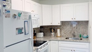 A compact kitchen with white cabinets, a stainless steel refrigerator, a microwave, a stove, and a sink. There are two teal mats on the floor, and a trash bin is visible. - Beachside Unit 29 - Key Colony (Key Colony Beach)