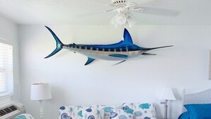 A room with a ceiling fan, a wall-mounted fish decoration, a patterned sofa, a white coffee table, and a bed with a blue cover. - Beachside Unit 29 - Key Colony (Key Colony Beach)