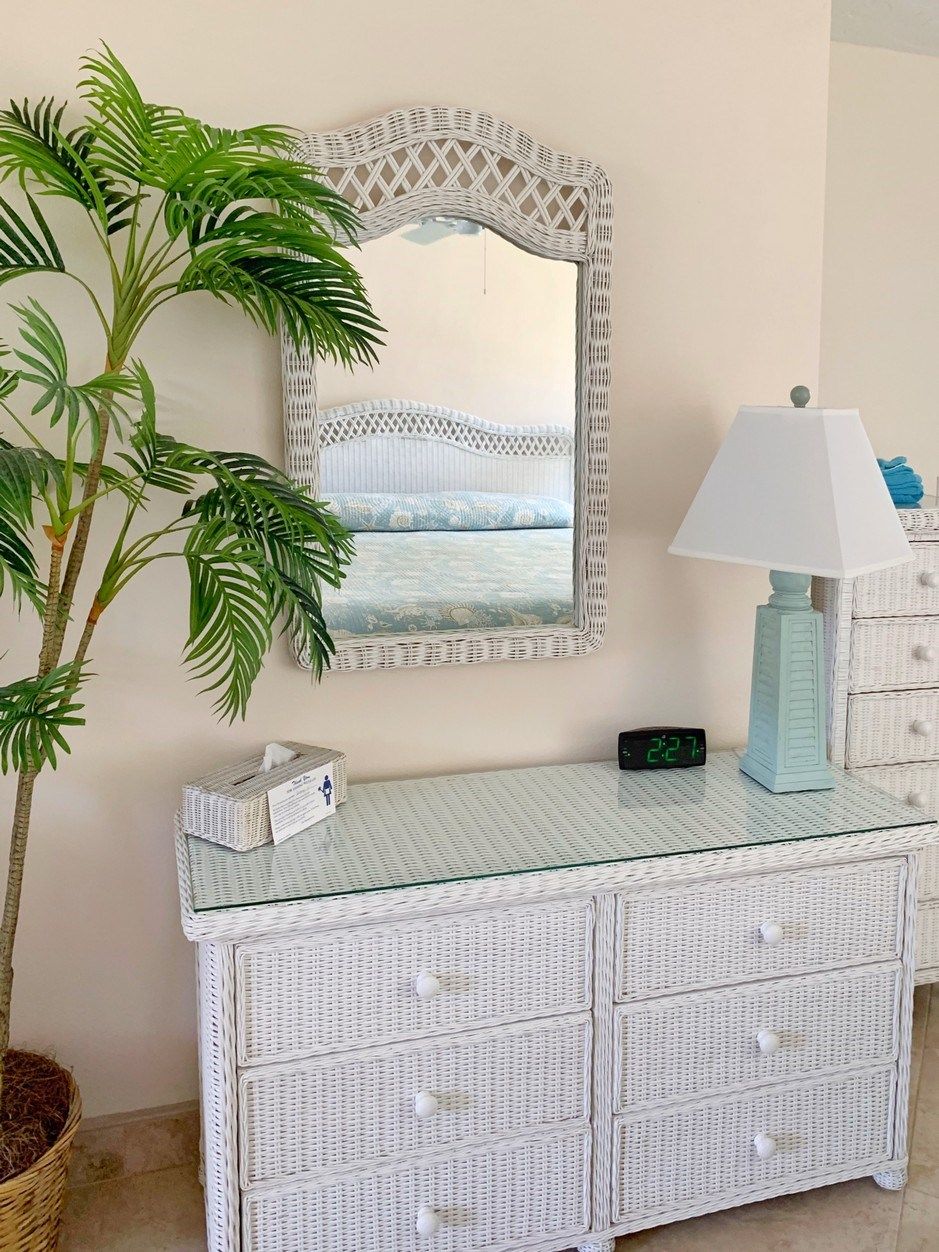 A room with a white wicker console table, a mirror with a lattice frame, a potted plant, a lamp, and a digital clock.