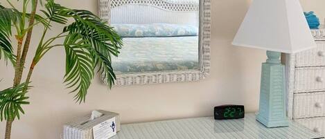 A room with a white wicker console table, a mirror with a lattice frame, a potted plant, a lamp, and a digital clock.