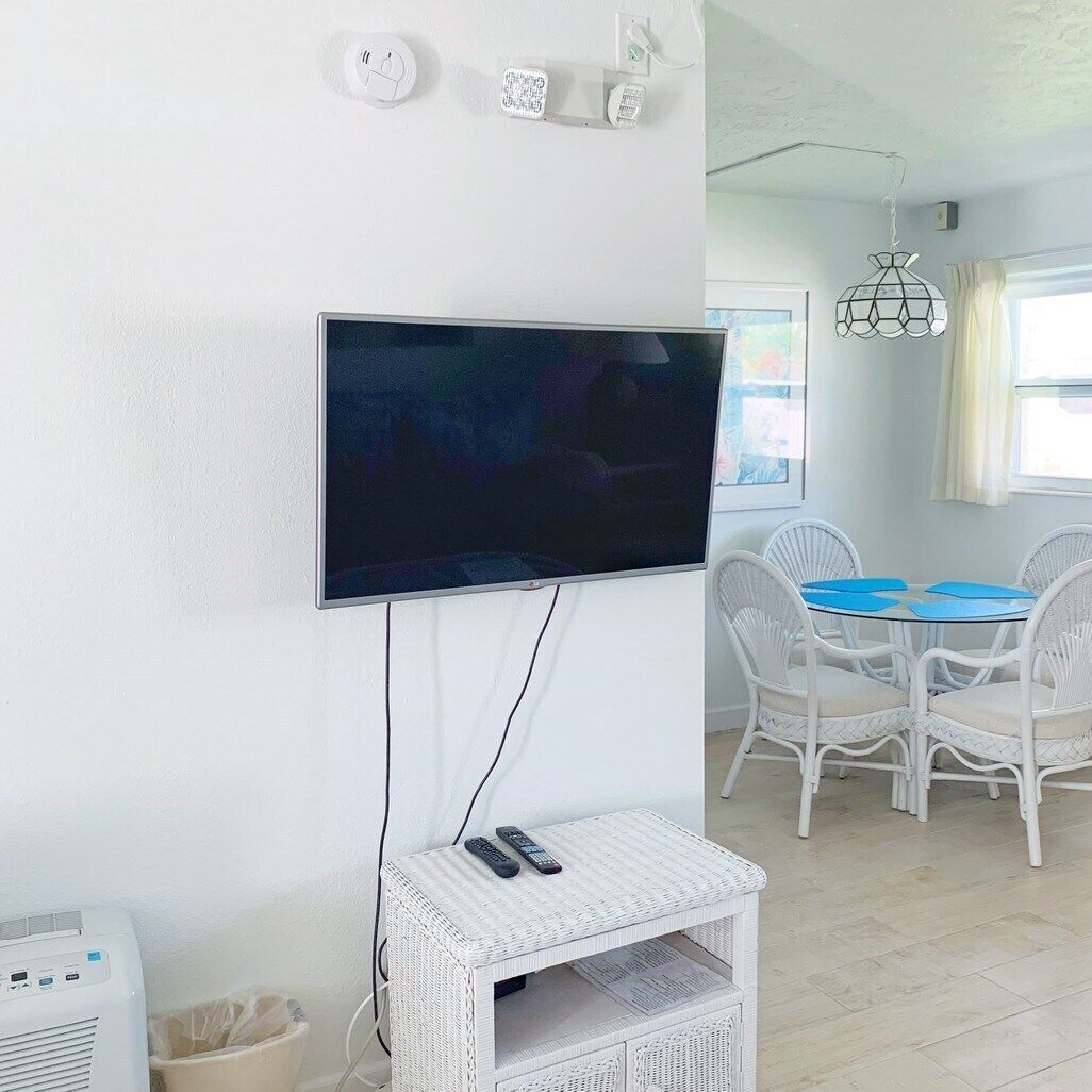Ein Zimmer mit einem wandmontierten Flachbildfernseher, einem weißen Rattan-Nachttisch mit Fernbedienungen, einer Essecke mit weißen Rattan-Stühlen und einem blauen Tisch sowie einer Deckenleuchte.
