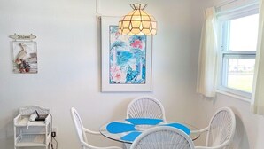 A dining area with a white wicker table and chairs, a hanging light fixture, a framed floral artwork, and a wicker shelf unit. - Beachside Unit 27 - Key Colony, Beachside Unit 27 - The Beach is Calling You! (Key Colony Beach)