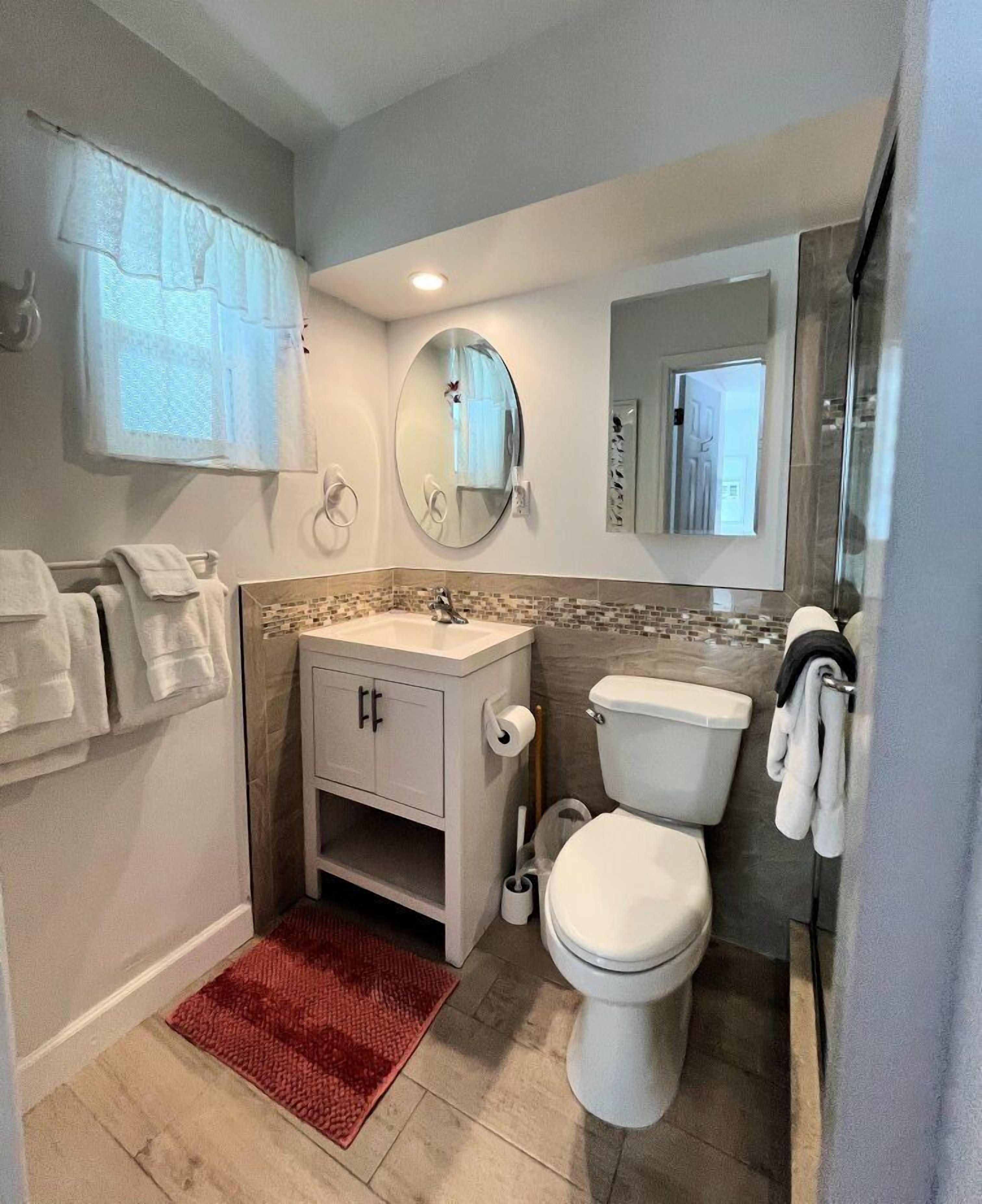 Ein Badezimmer mit Toilette, Waschbecken und Spiegel. Handtücher hängen an der Wand und ein roter Teppich liegt auf dem Boden.