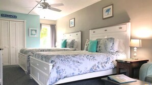 A bedroom with two beds, a ceiling fan, a nightstand with a lamp, and a desk with a chair.