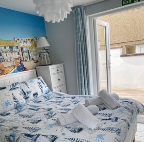A bedroom with a bed featuring a nautical-themed bedspread, a white bedside table, a lamp, and a view of a coastal street through a window. - Gull Rock is a Frontline House above Porthmeor Beach, with Amazing Sea Views (St Ives)