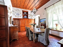 Un comedor con una mesa de madera, sillas y una araña. Hay un televisor en la pared y una ventana con cortinas.