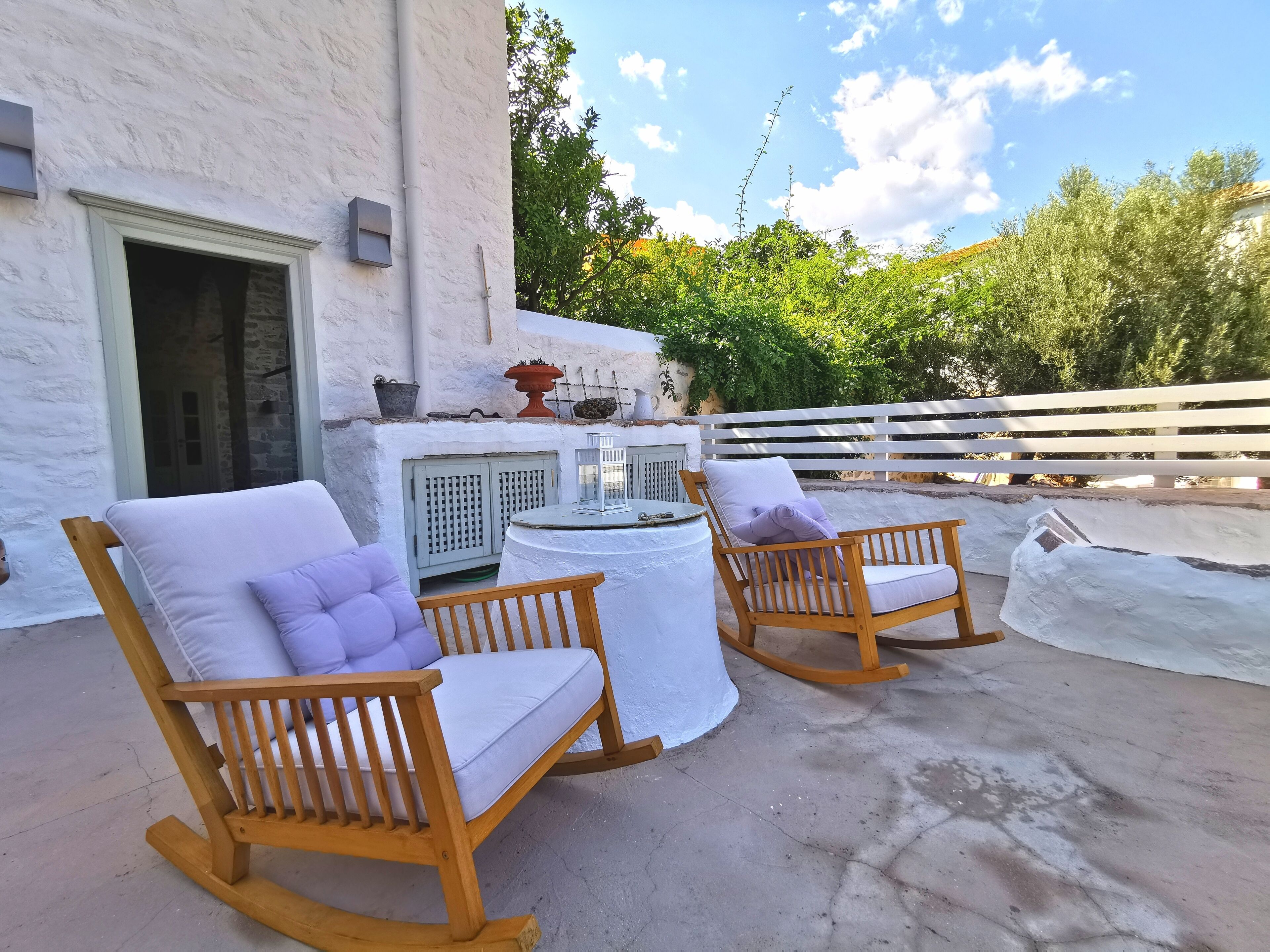 A patio with two rocking chairs and a small table, a white wall, and a wooden fence in the background.