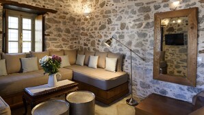 A stone-walled room with a sofa, a coffee table with a vase, a lamp, and a television. - Cotommatae Hydra 1810 (Hydra)