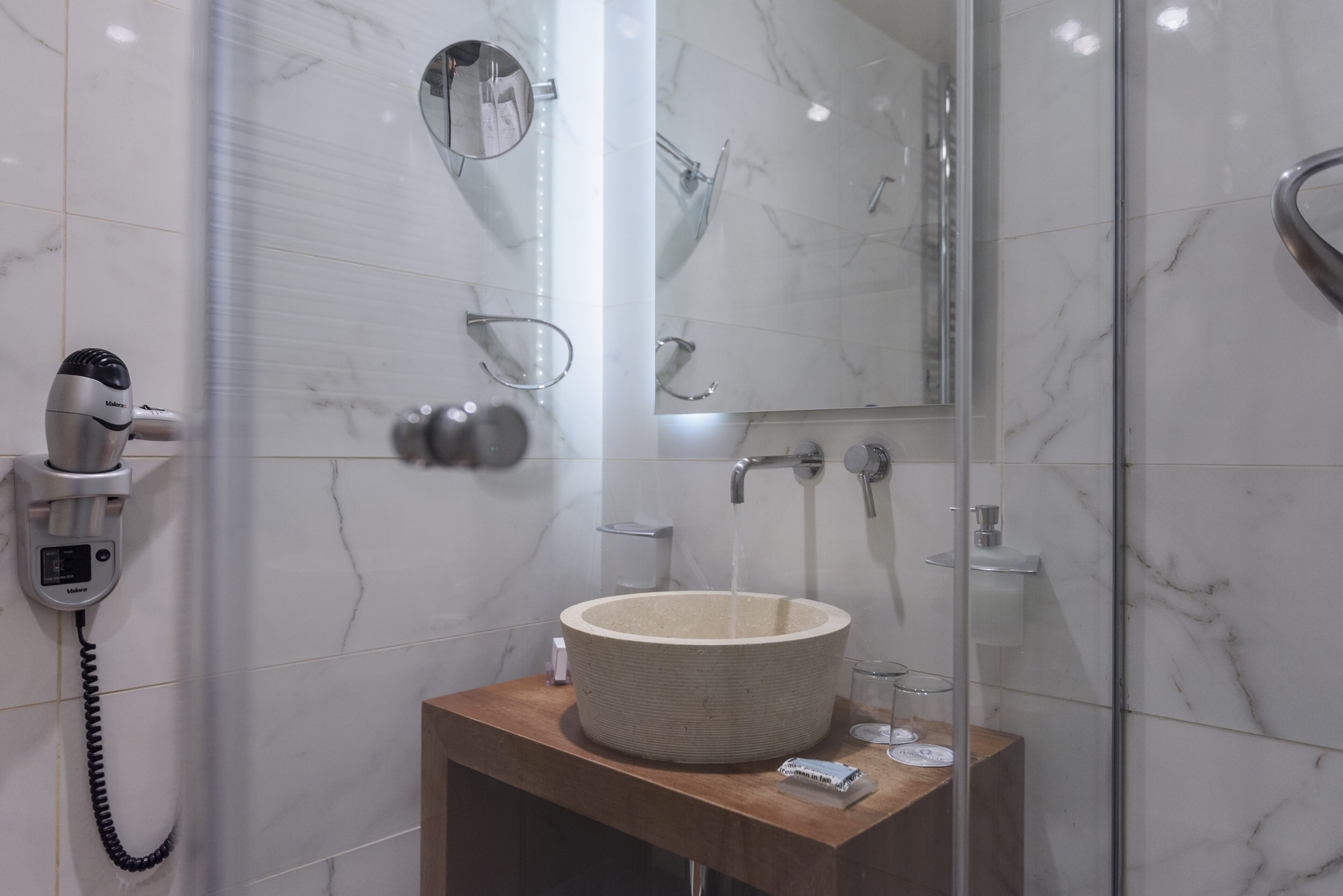 Ein Badezimmer mit einer marmorverkleideten Wand, einem hölzernen Waschtisch, einem großzügigen Waschbecken, einer Handbrause, einer Duschkabine und einem an der Wand montierten Fön.