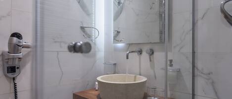 Ein Badezimmer mit einer marmorverkleideten Wand, einem hölzernen Waschtisch, einem großzügigen Waschbecken, einer Handbrause, einer Duschkabine und einem an der Wand montierten Fön.