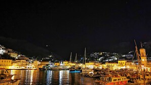 A nighttime harbor with boats docked, a clock tower, and buildings along the waterfront. - Cotommatae Hydra 1810 (Hydra)
