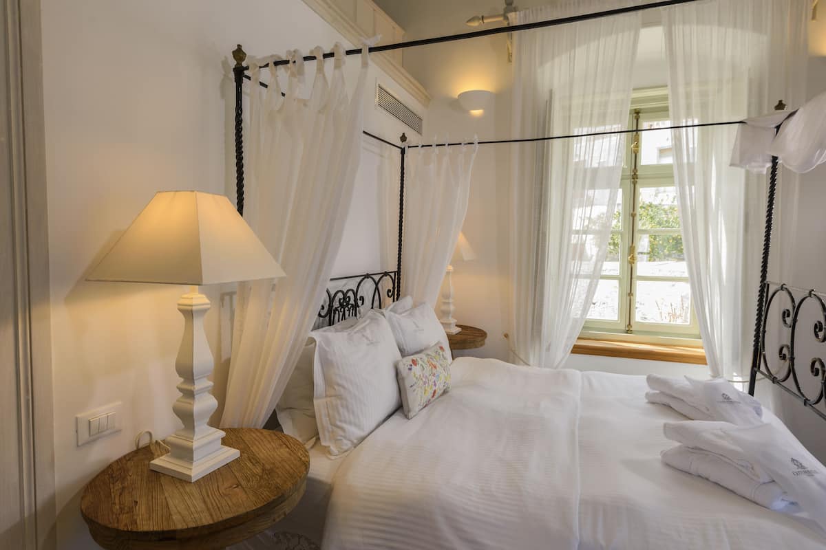 A four-poster bed with white linens, a bedside table with a lamp, and sheer curtains.