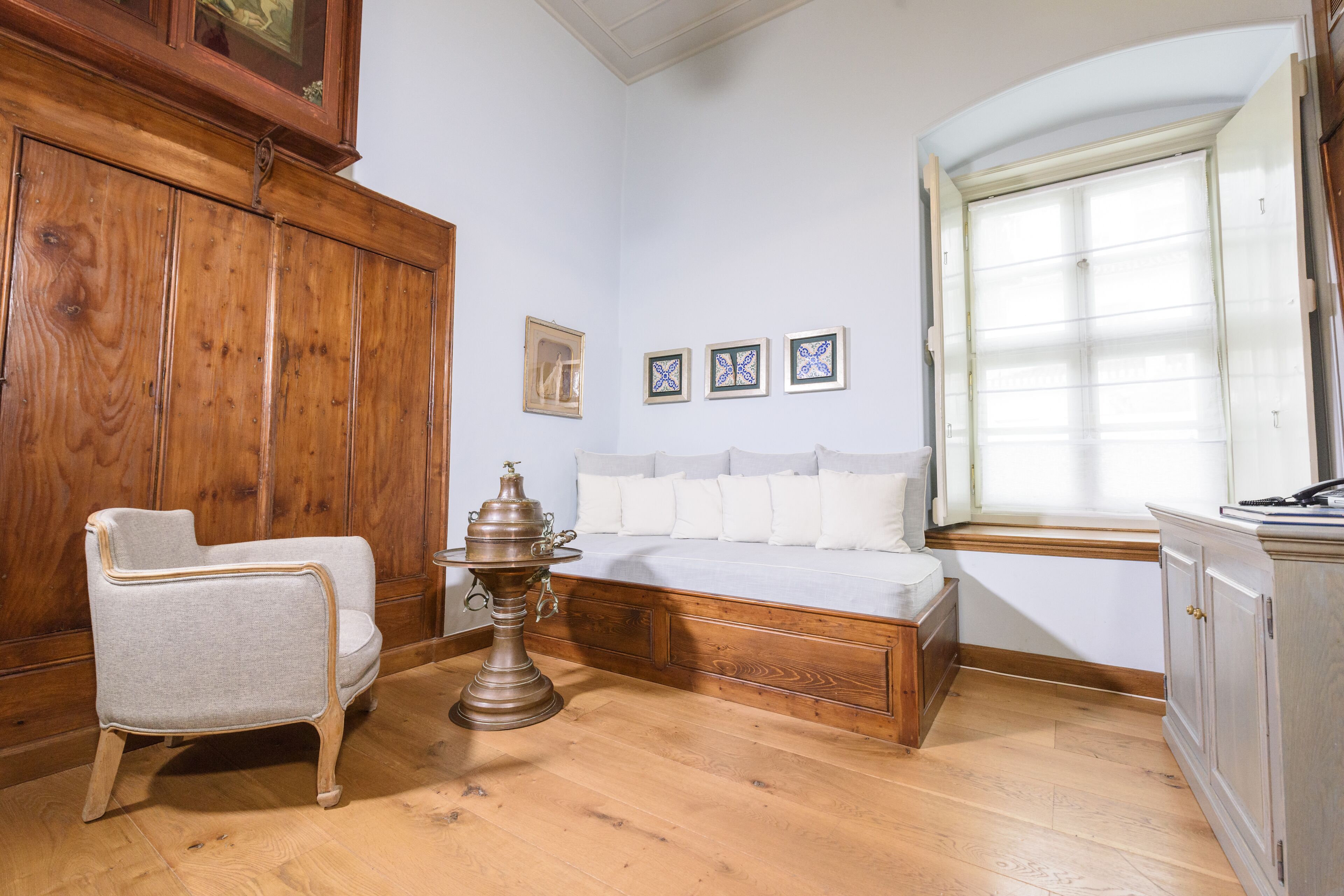 Una habitación con un armario de madera, un sillón blanco, un banco de madera y una ventana con persianas.
