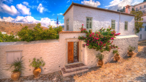 A white building with a wooden door, potted plants, and a cobblestone street. - Cotommatae Hydra 1810 (Hydra)
