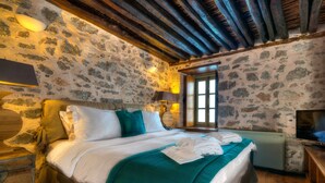 A stone-walled bedroom with a wooden ceiling, a bed with white and teal bedding, a television, and a window with a view. - Cotommatae Hydra 1810 (Hydra)