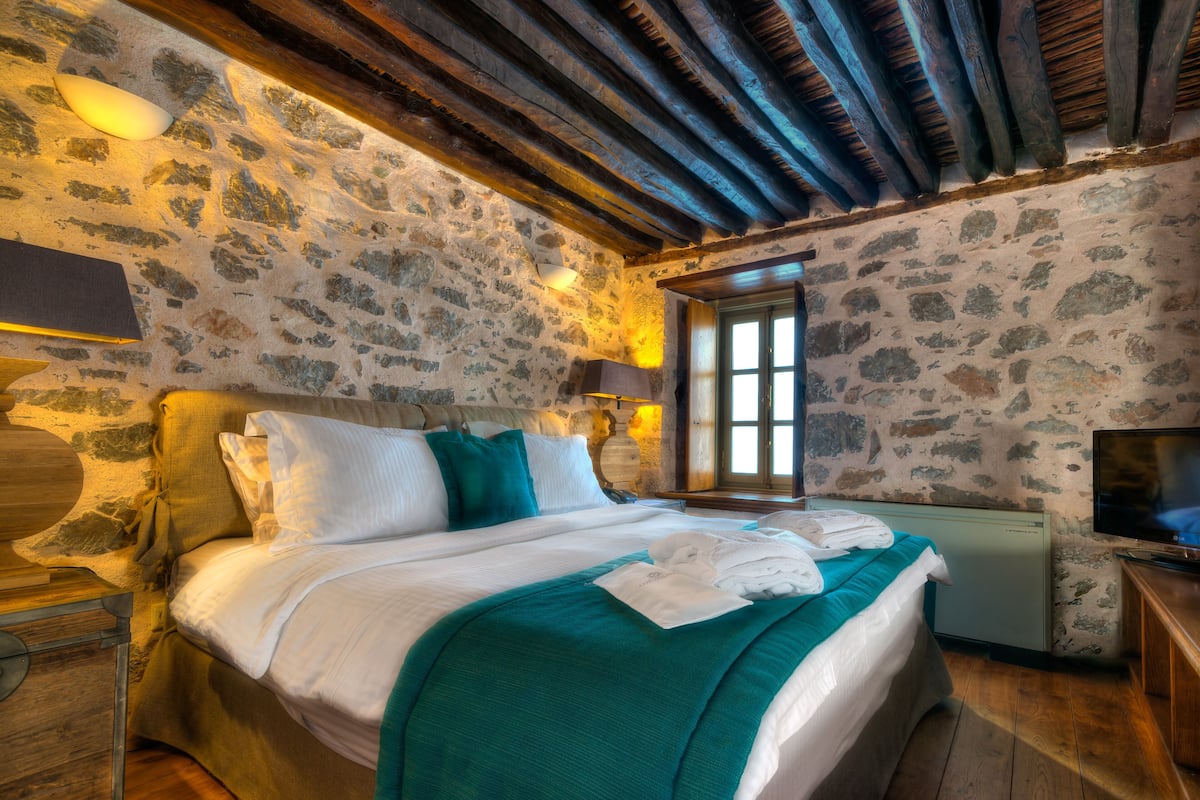 A stone-walled bedroom with a wooden ceiling, a bed with white and teal bedding, a television, and a window with a view.