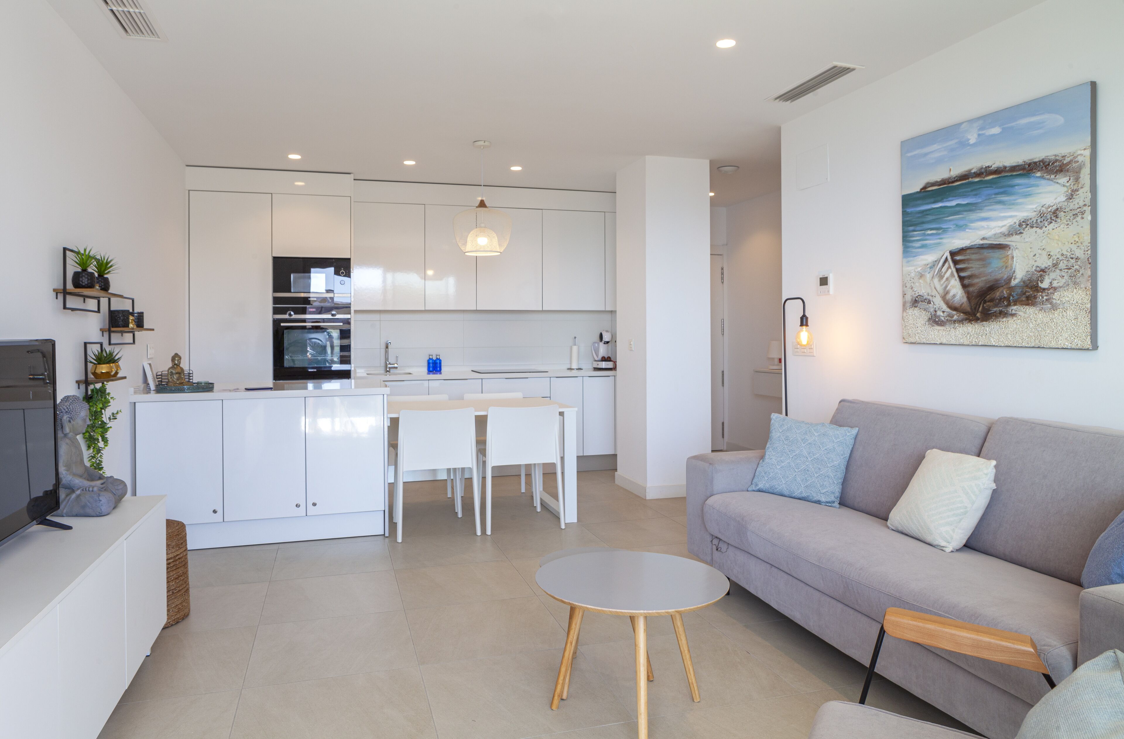 Une cuisine moderne avec des meubles blancs, un coin repas avec une table et des chaises, un canapé gris avec des coussins et un tableau accroché au mur.
