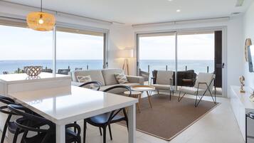 A modern living room with a dining area, large windows, and a view of the sea.